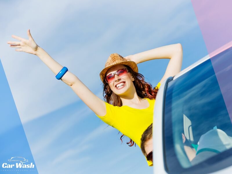 A joyful woman with red sunglasses and a straw hat sticks her head out from a car window on a sunny day, her arms raised in happiness after a satisfying car wash experience, with the Car Wash Near Me logo featured in the corner.